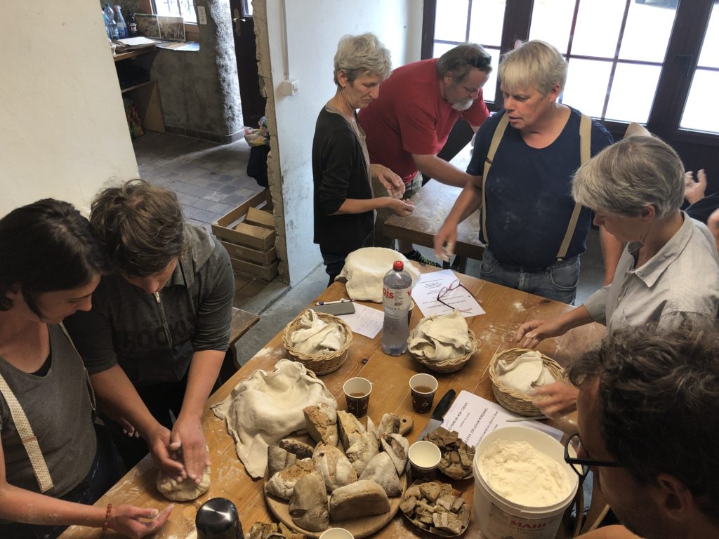 Membres de l'association pétrissant et façonnant de la pâte à pain autour d'une grande table, avec des paniers, des pains au levain et des supports de cours.