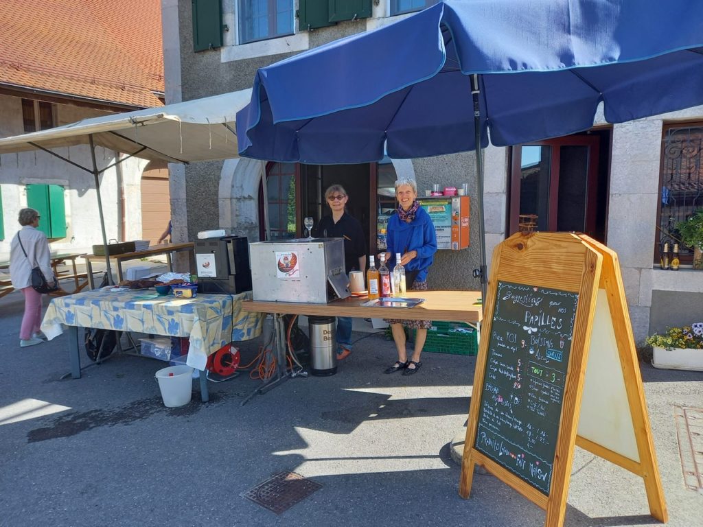 Deux bénévoles souriantes derrière un stand de boissons et de nourriture sous un grand parasol bleu, à côté d'une ardoise annonçant les pizzas et produits faits maison.