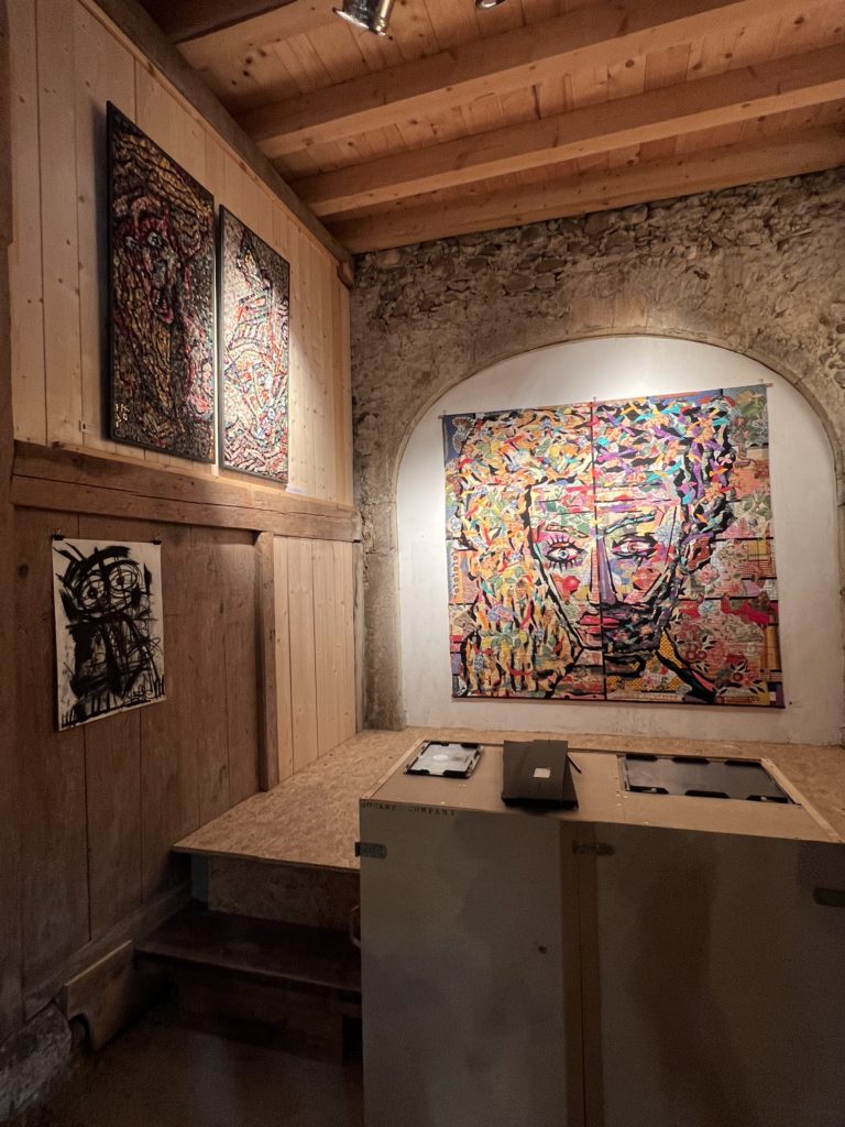 Coin de la Grange aménagé en galerie : un grand portrait en collage très coloré sous une voûte de pierre éclairée, deux tableaux colorés et un dessin au fusain sur les murs en bois.