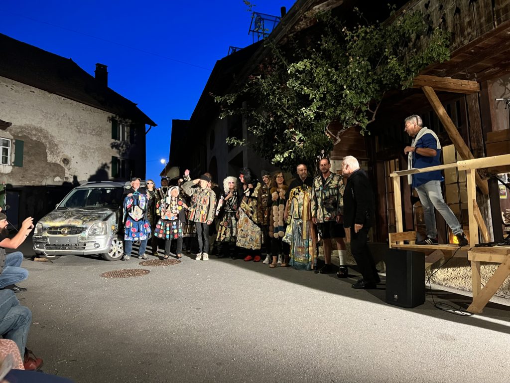 À la tombée de la nuit, une vingtaine d'habitants alignés dans une rue du village portent des vêtements colorés et décorés, à côté d'une voiture entièrement peinte, devant un public assis.