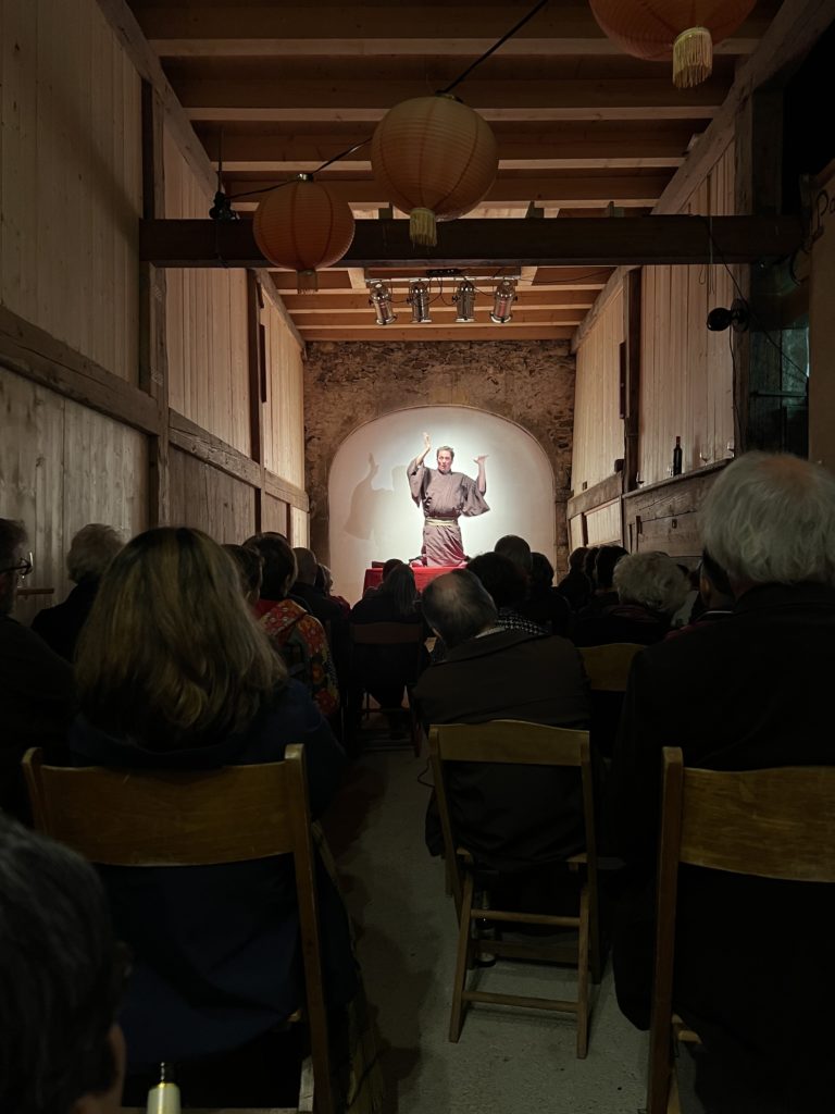 Vue depuis le fond de la Grange : le public assis sur des chaises en bois, sous des lanternes en papier orange, regarde le conteur en kimono sur une petite scène éclairée devant un mur de pierre.