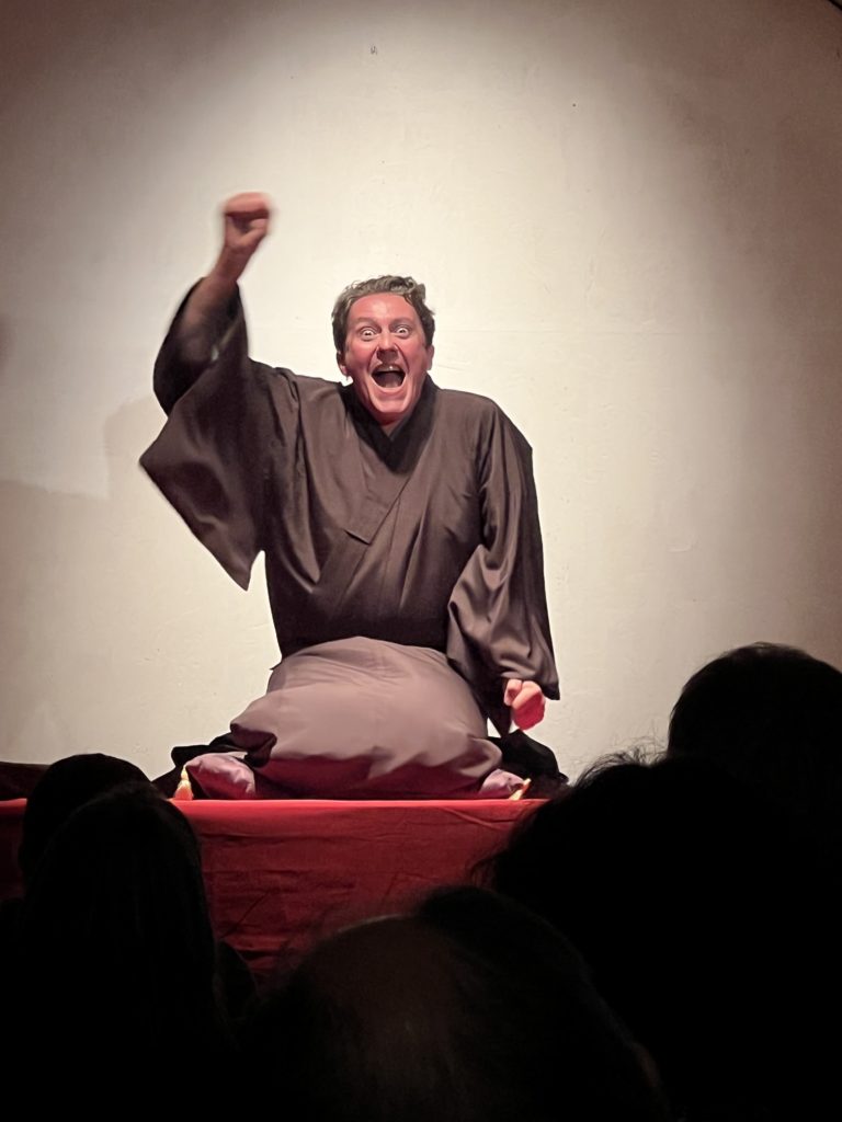 Le conteur Stéphane Ferrandez, en kimono, à genoux sur un coussin posé sur une estrade rouge, le poing levé et le visage très expressif, devant le public vu de dos.
